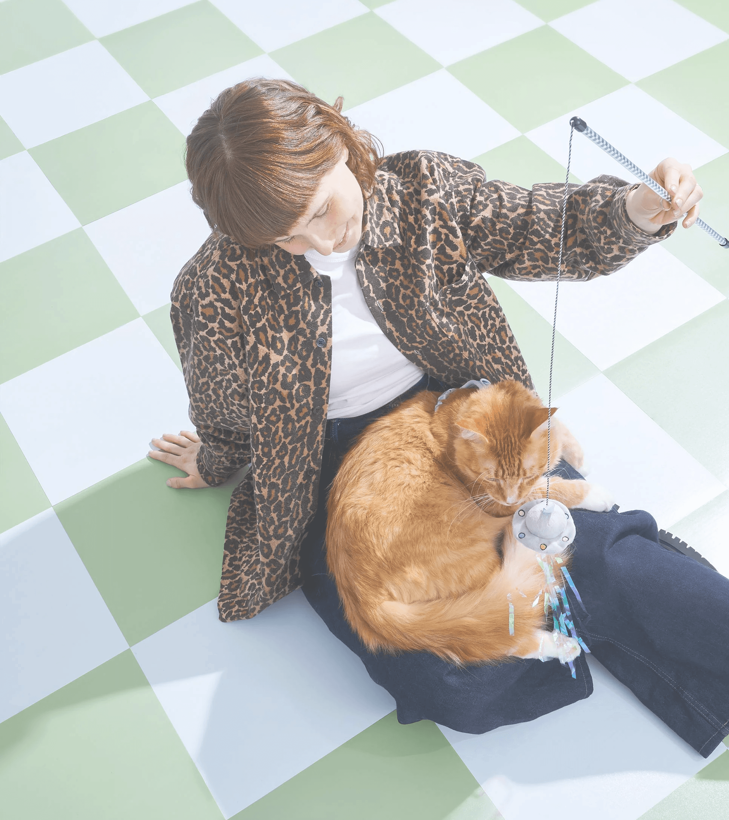 A pet parent sitting on a checkerboard floor, teasing a ginger cat curled in their lap with a wand toy
